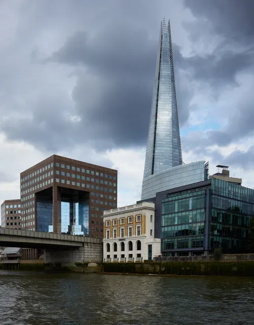 This urban architecture photograph captures the London skyline in the late afternoon during the summer. The main subject of the image is The Shard, a prominent glass skyscraper in central London, which stands tall against dramatic summer clouds. In the foreground, the River Thames flows beside notable riverside buildings, including a historic structure and modern office complexes, with the characteristic city architecture well represented. The photograph provides a clear view of The Shard as a landmark, illustrating its imposing presence along the Thames and highlighting the contrast between old and new architectural styles that define this part of London.